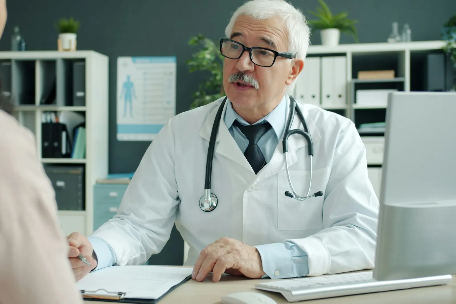 A physician in a white coat talking with an adult patient across a desk in a medical office