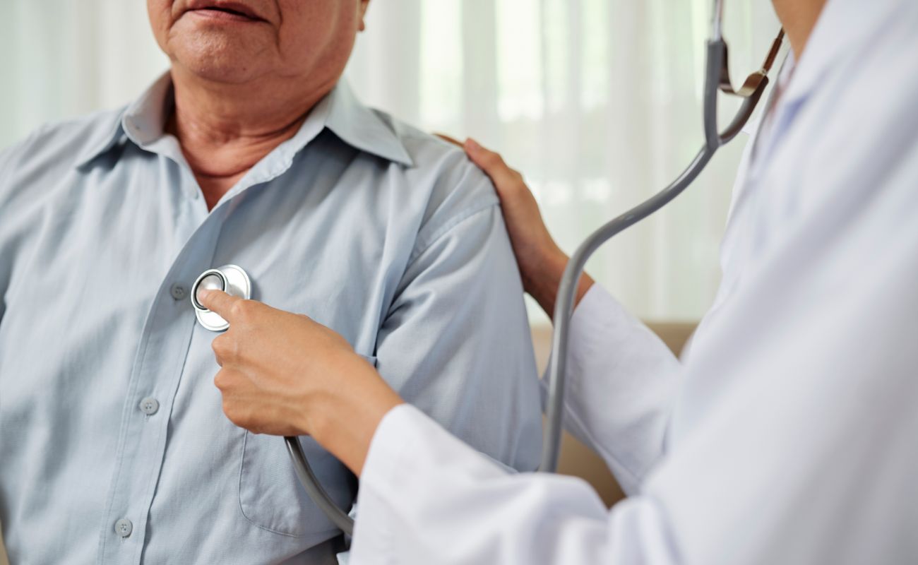 A doctor uses a stethoscope to listen to a patient's heartbeat. The patient is sitting and wearing a light blue shirt.