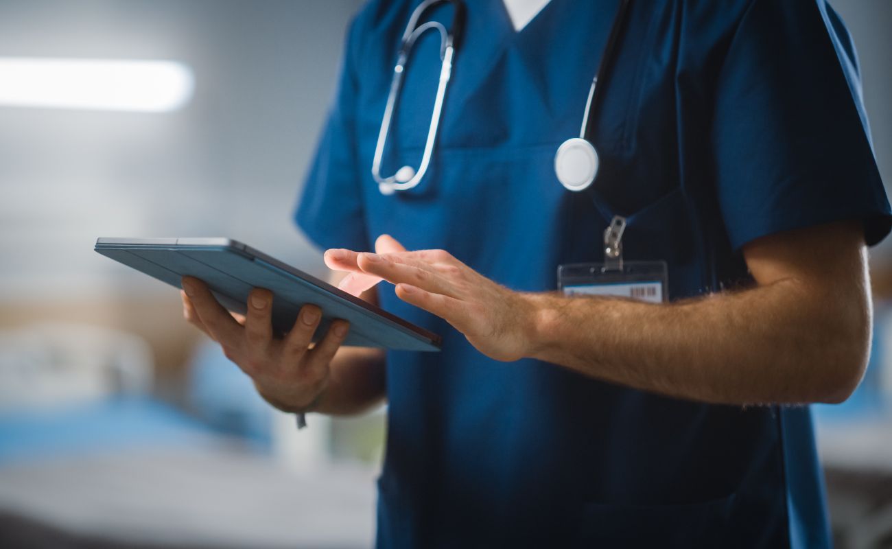 A healthcare professional in blue scrubs uses a tablet device, with a stethoscope around their neck, in a medical setting.