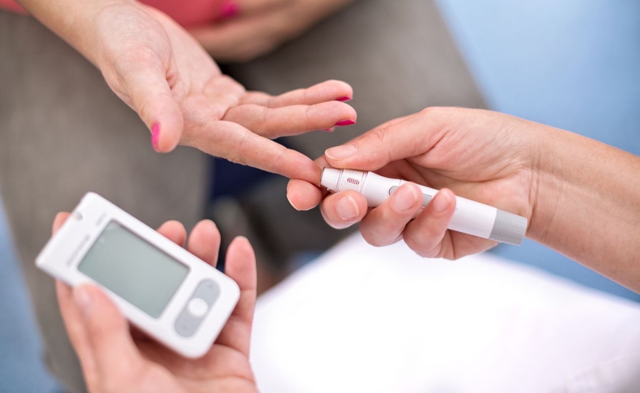 A person uses a lancet to prick another person's fingertip. A glucometer is visible, possibly indicating a blood sugar test.